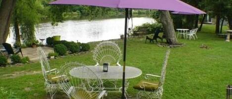 A round table with four white chairs and a purple umbrella in a grassy area near a body of water.