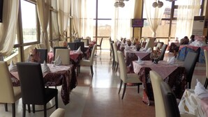 A restaurant with tables covered in patterned tablecloths, chairs, and a large window with curtains. - Hotel des Deputes (Yaoundé)