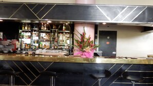 A bar area with a curved counter, multiple shelves of bottles, and a pink floral arrangement. - Hotel des Deputes (Yaoundé)