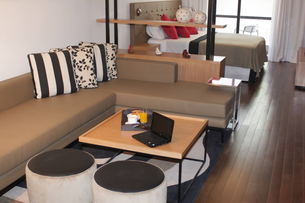 A modern hotel room with a sofa, a small table with a laptop, a bed with a headboard, and a window with curtains.