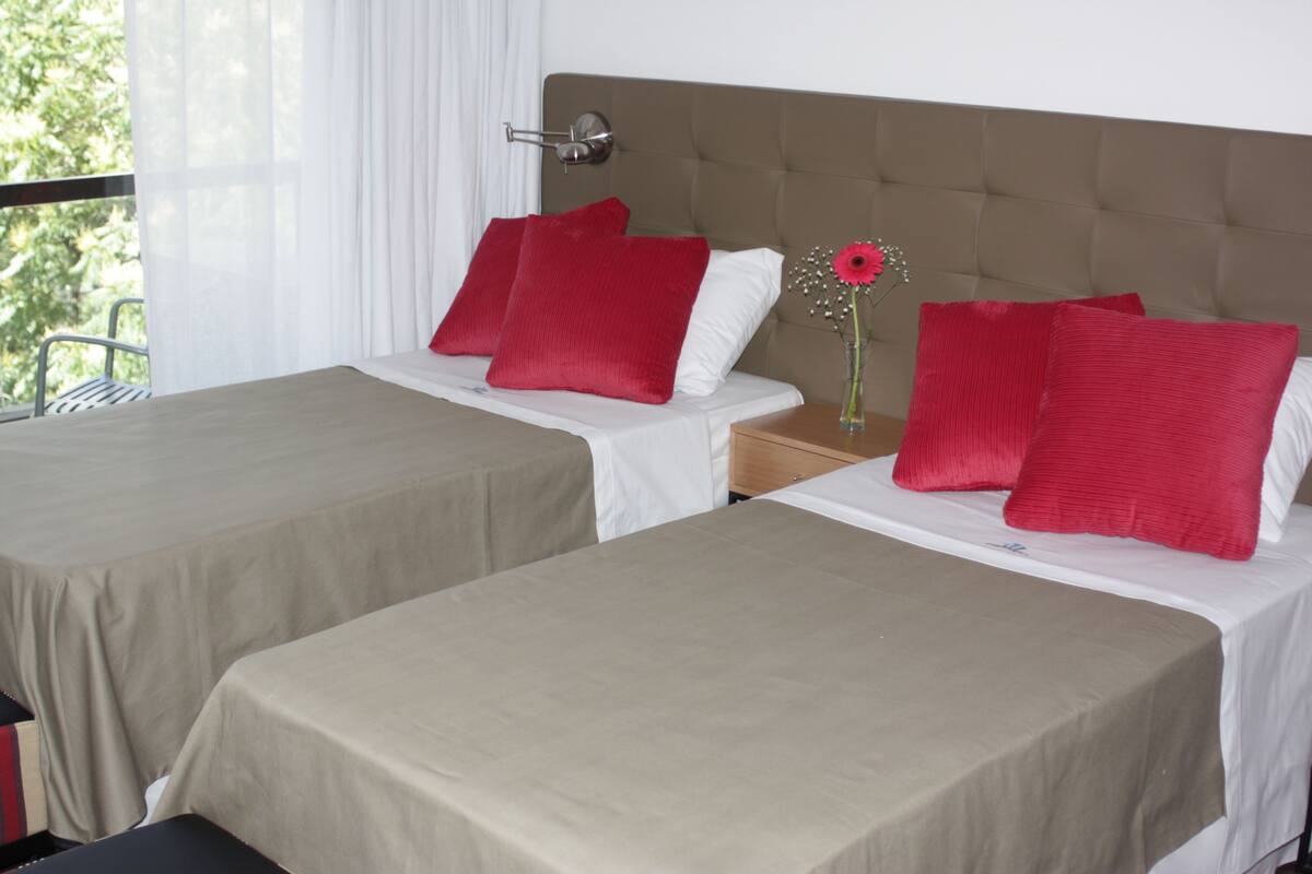 Two single beds with red and white pillows, a wooden nightstand, and a vase with a flower.