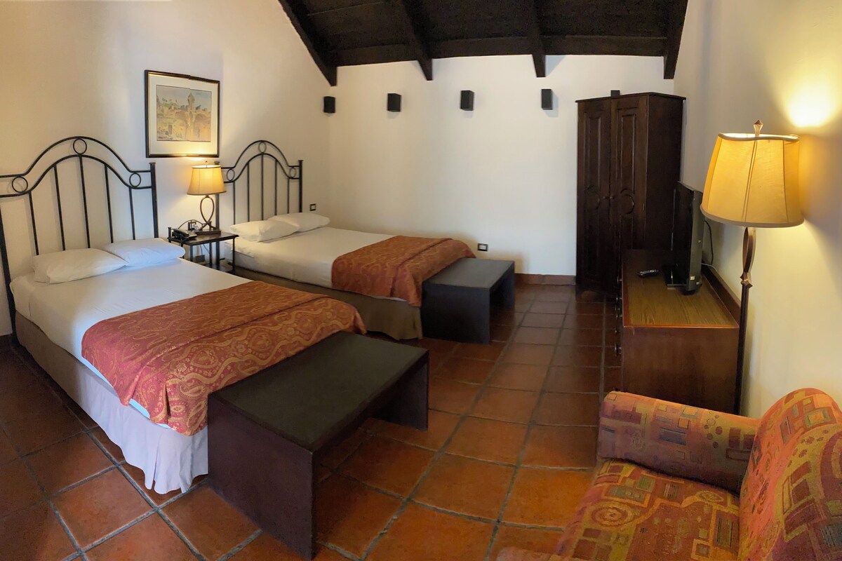 Una habitación con dos camas, un banco de madera, una silla, un armario de madera, un cuadro en la pared y una lámpara.