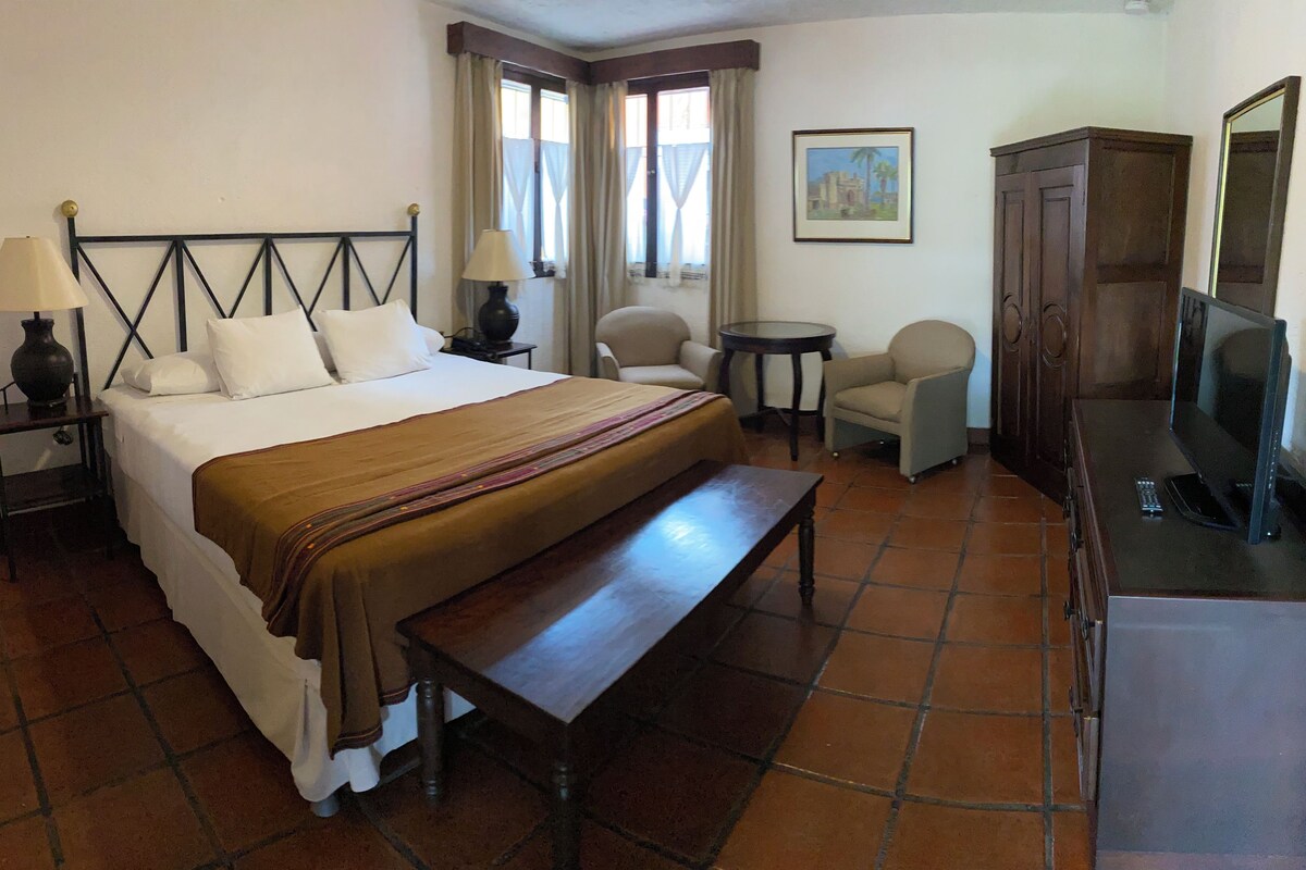Habitación de hotel con cama, un banco, escritorio, silla, televisor y un cuadro en la pared.