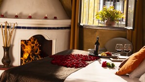 A room with a fireplace, a bed with rose petals, a tray of fruit, wine glasses, and a potted plant by the window. - Villa Colonial (Antigua Guatemala)