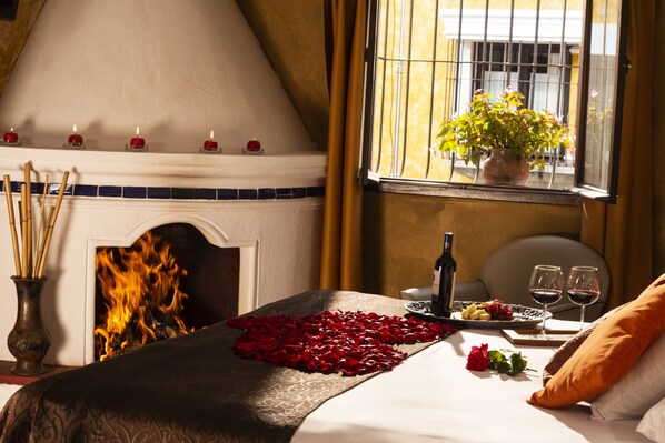 A room with a fireplace, a bed with rose petals, a tray of fruit, wine glasses, and a potted plant by the window.