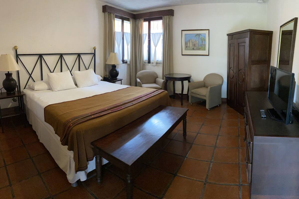 A hotel room with a bed, a bench, a desk, a chair, a television, and a painting on the wall.