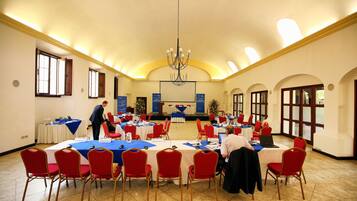Une salle de conférence équipée de tables et de chaises, d’un lustre et d’une scène avec un pupitre.