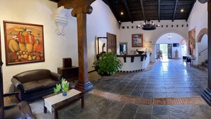 A spacious lobby with a reception desk, a seating area with a sofa and a table, a large painting on the wall, and a tiled floor. - Villa Colonial (Antigua Guatemala)