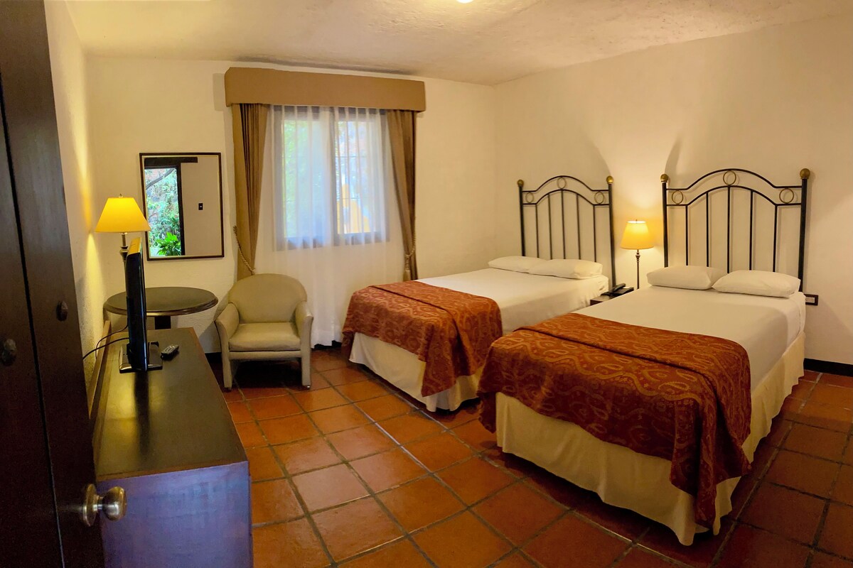Habitación de hotel con dos camas, un escritorio, una silla y una ventana con cortinas.