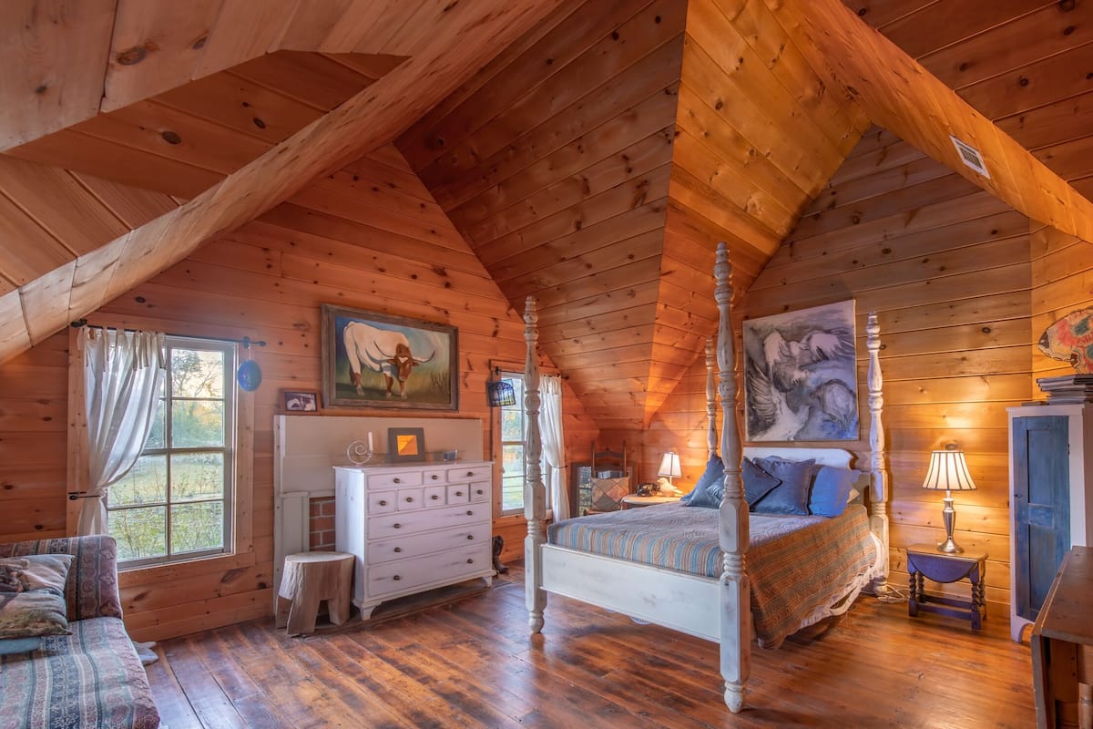 A cozy log cabin bedroom with a four-poster bed, a dresser, a small table, and a window with curtains.