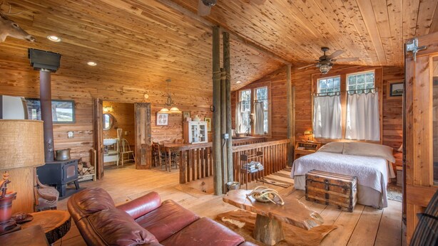 A log cabin interior with a wooden floor, a dining area, a kitchen with a wood stove, a bedroom with a bed and a window, and a bathroom with a sink and mirror. - Seventy-four Ranch (Jasper)