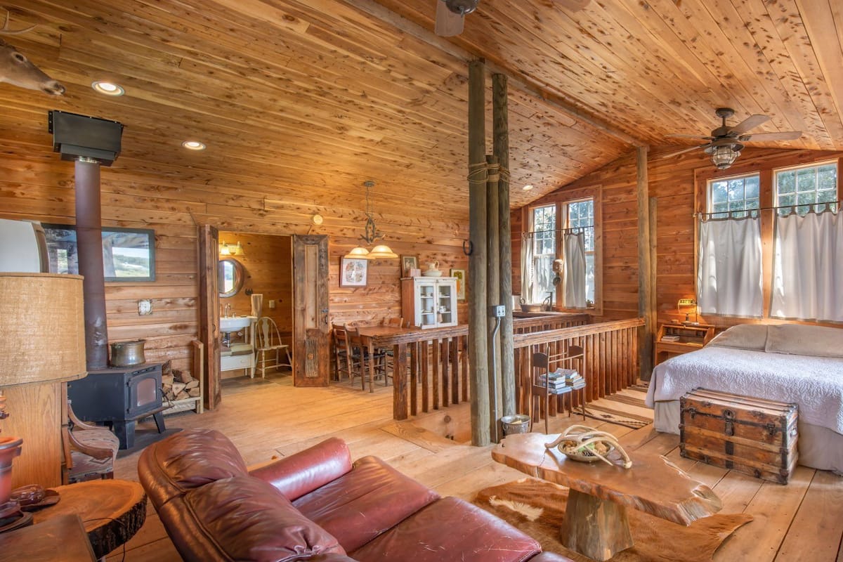 A log cabin interior with a wooden floor, a dining area, a kitchen with a wood stove, a bedroom with a bed and a window, and a bathroom with a sink and mirror.
