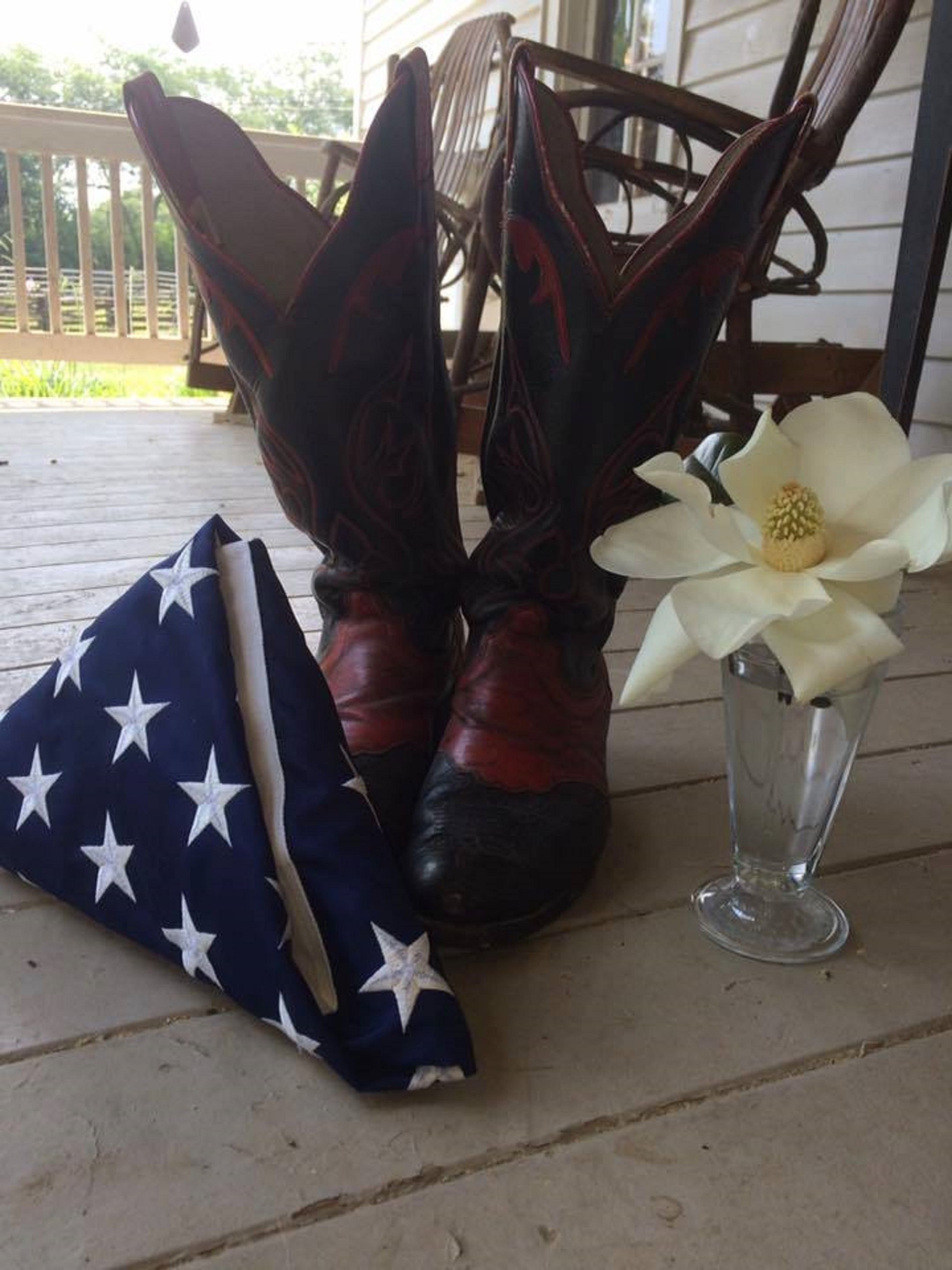 A pair of cowboy boots, a small American flag pillow, and a white flower in a glass vase on a porch.