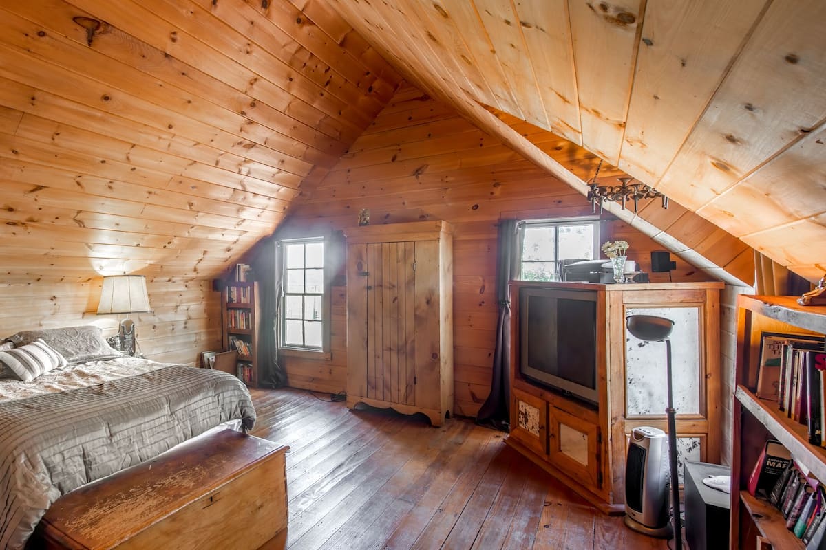 A cozy attic bedroom with a wooden ceiling, a bed with a patterned bedspread, a nightstand, a bookshelf, and a television.