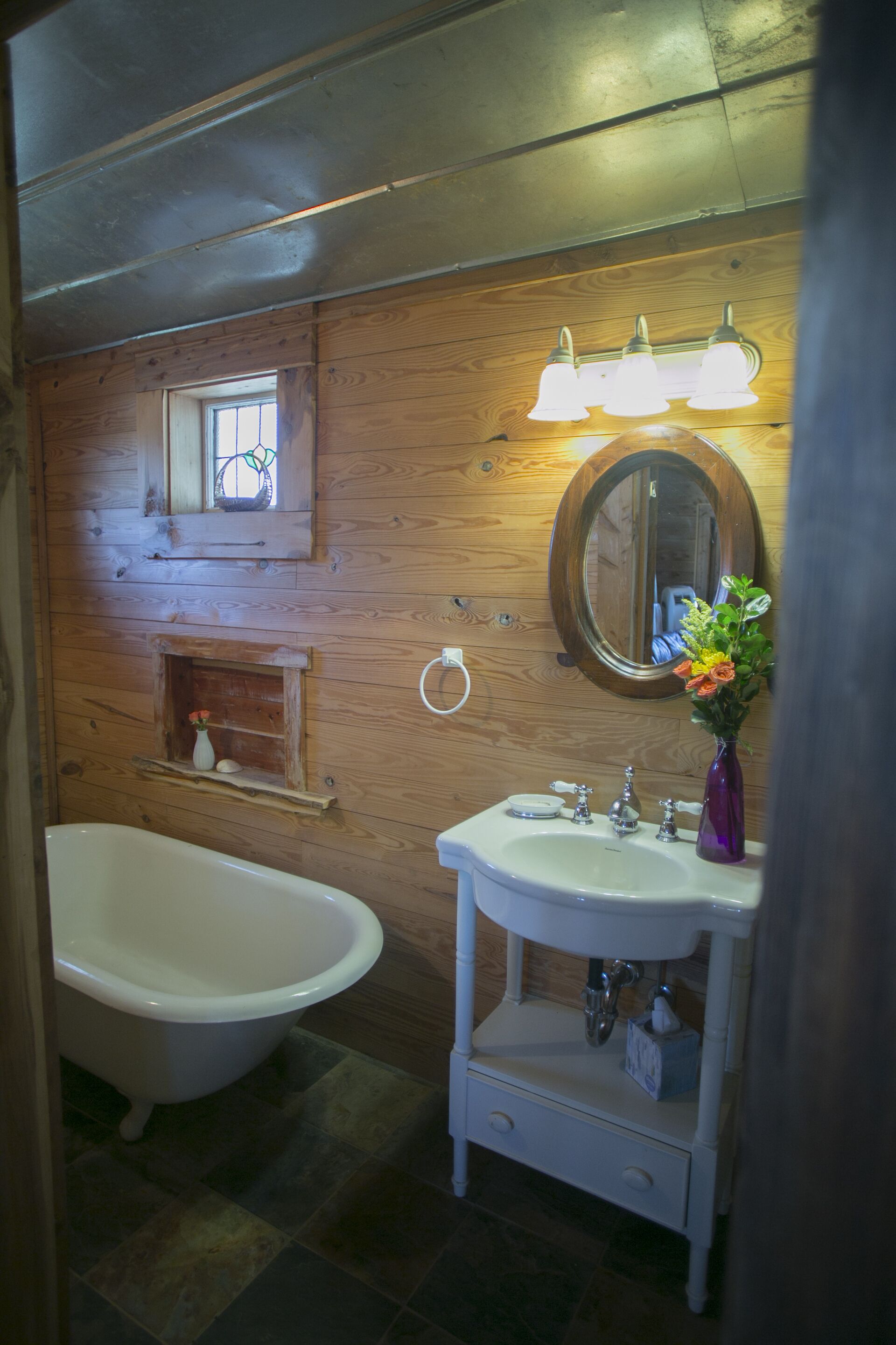 Un baño con pared de madera, una bañera independiente, un lavamanos con espejo y un florero con flores.