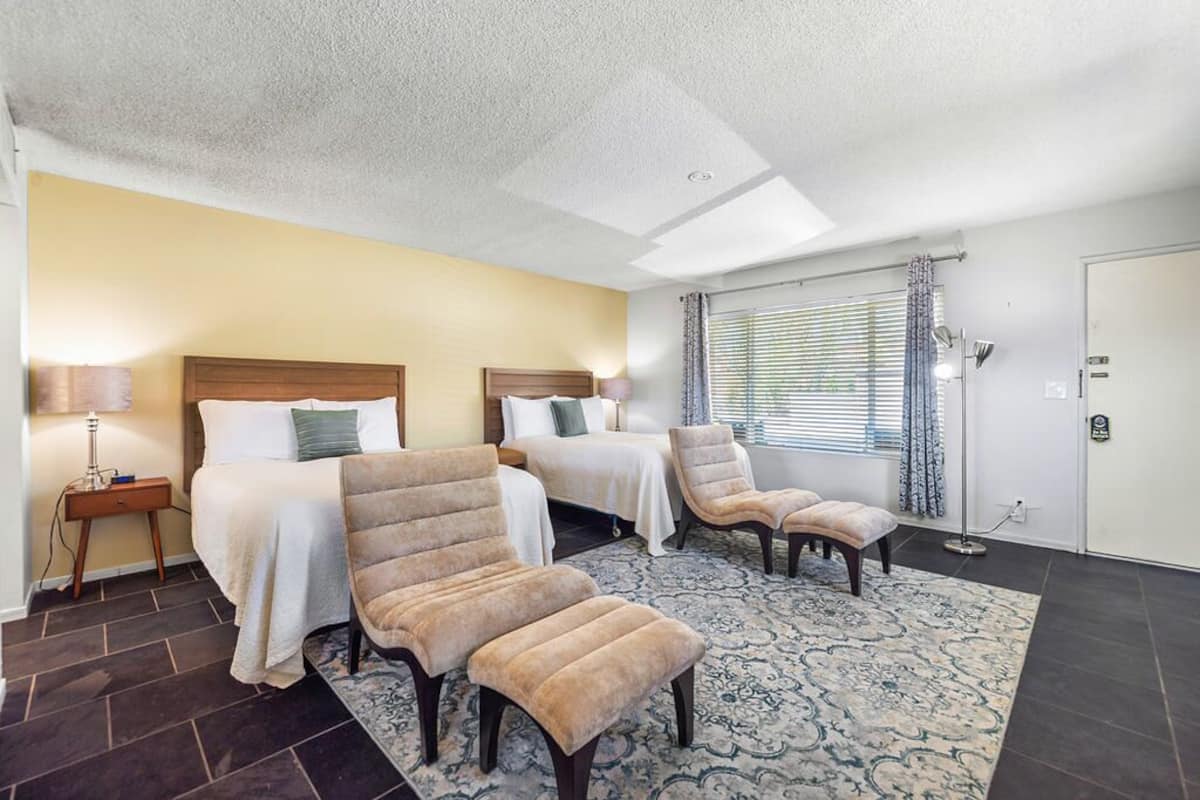 A hotel room with two beds, a sitting area with chairs, a patterned rug, and a window with curtains.