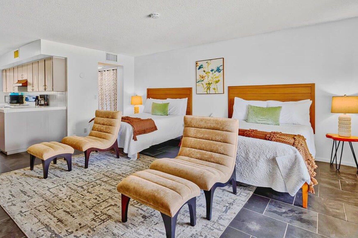 Habitación de hotel con dos camas, kitchenette, zona de estar con sillas y un tapete, y una mesita de noche con lámpara.
