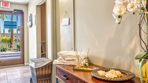 Zona de recepción de hotel con un mostrador de madera, un espejo y un jarrón con flores blancas. Hay una pila de toallas y una bandeja con aperitivos.