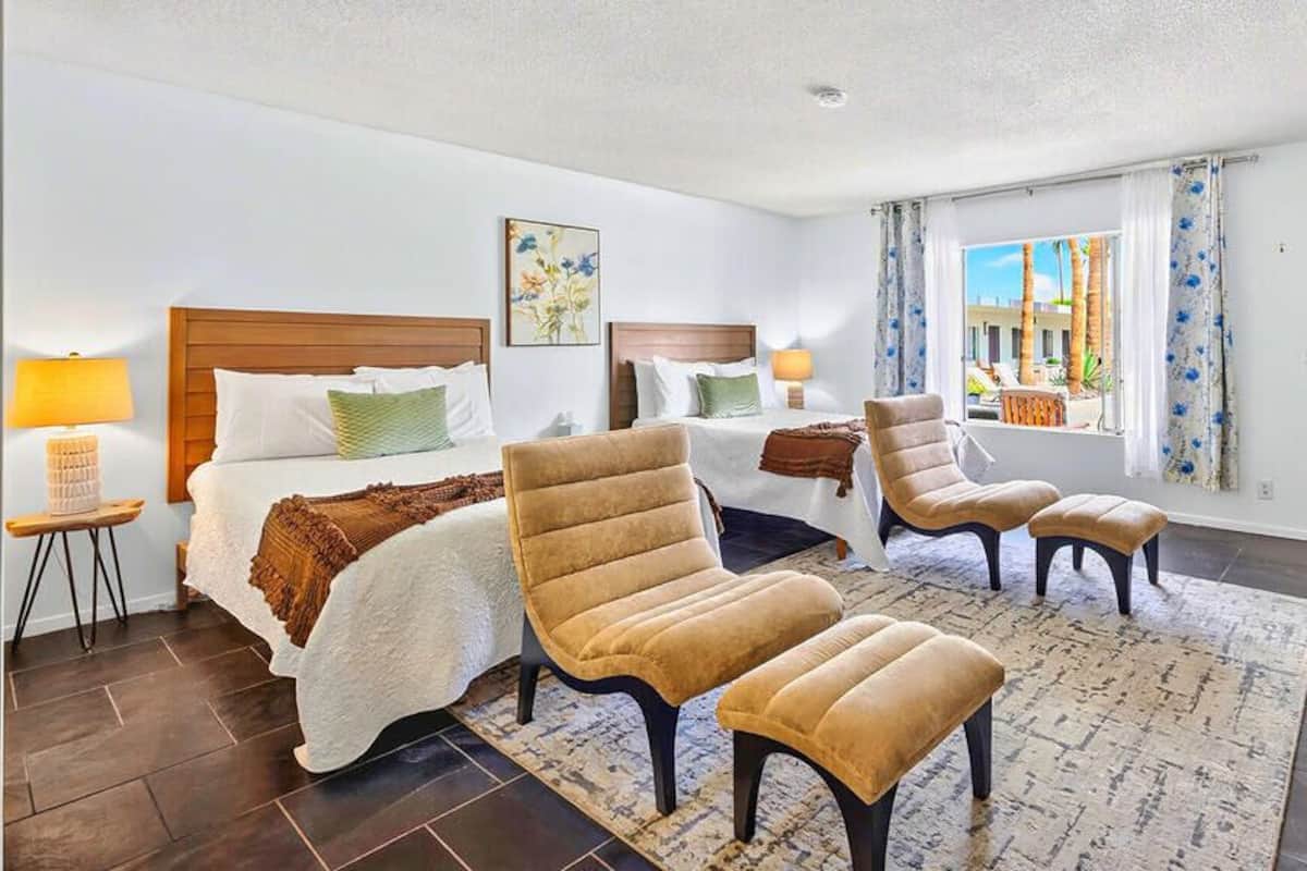 A hotel room with a bed, two armchairs, a small table, a window with curtains, and a view of buildings outside.
