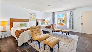 A hotel room with a bed, two armchairs, a small table, a window with curtains, and a view of buildings outside.