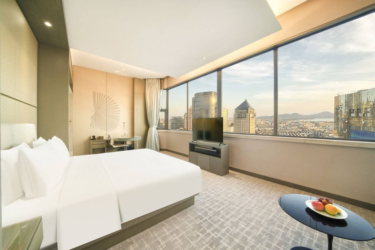 A hotel room with a large bed, a TV, a desk, and a view of the cityscape.