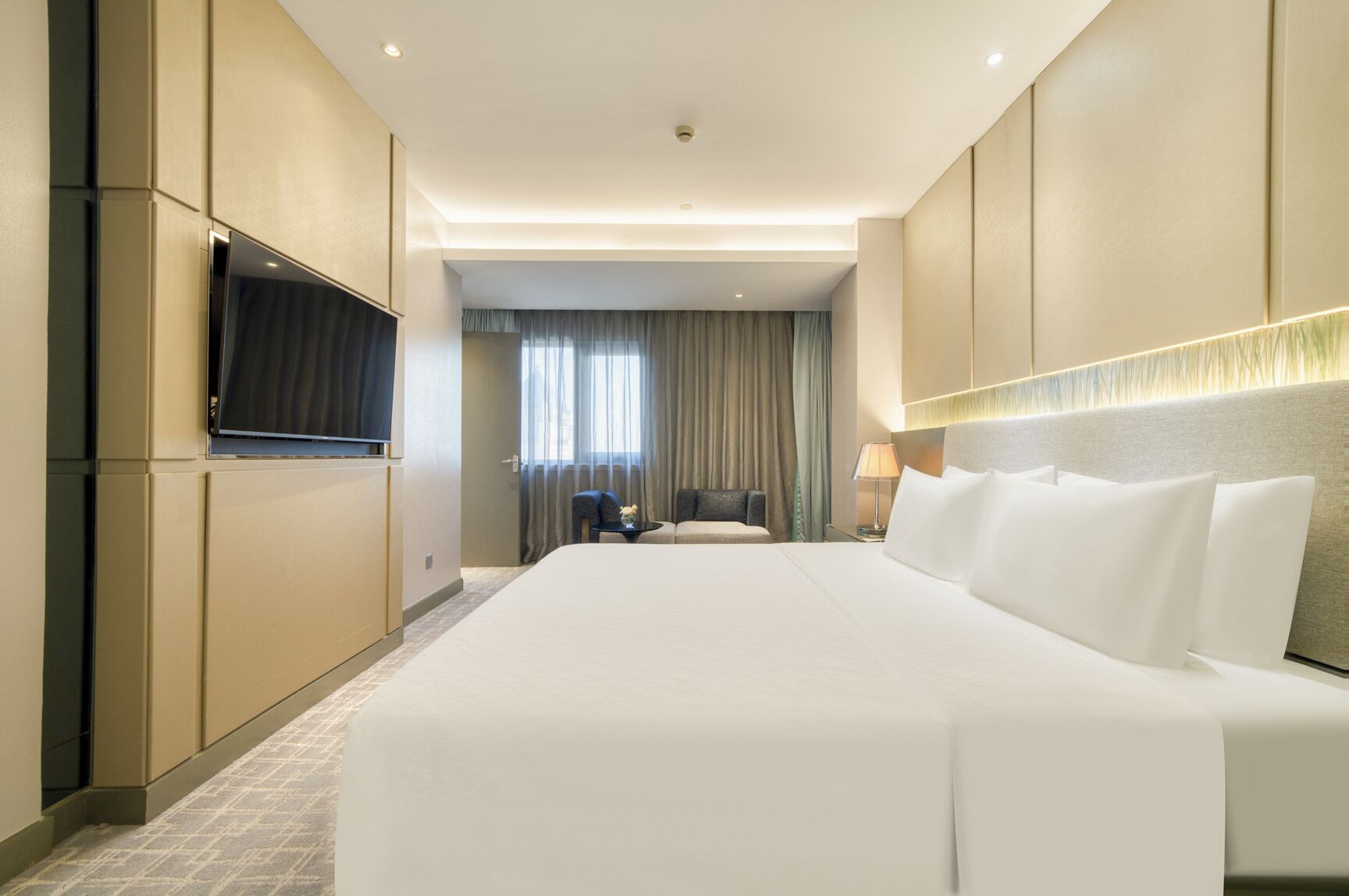 Habitación de hotel con una cama grande, un televisor de pantalla plana montado en la pared, una mesita de noche con lámpara y vistas al exterior a través de una ventana con cortinas.