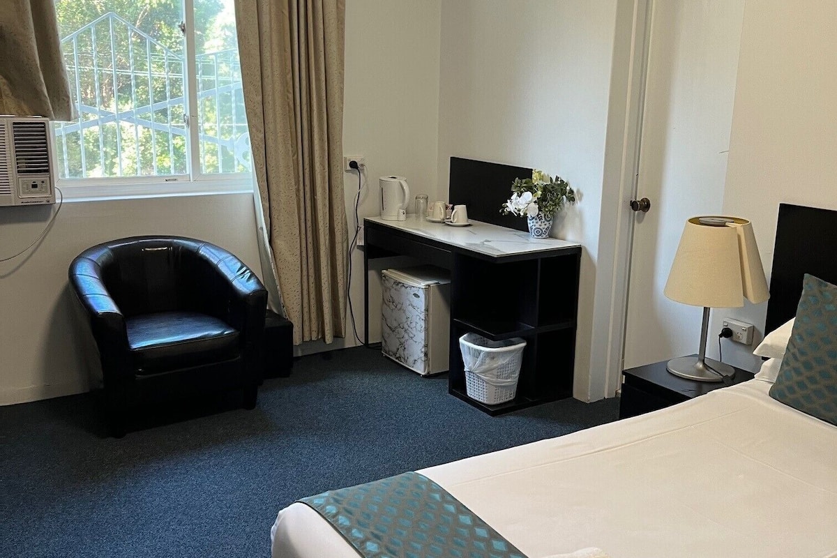 A hotel room with a bed, a desk, a chair, a window with curtains, and a lamp.
