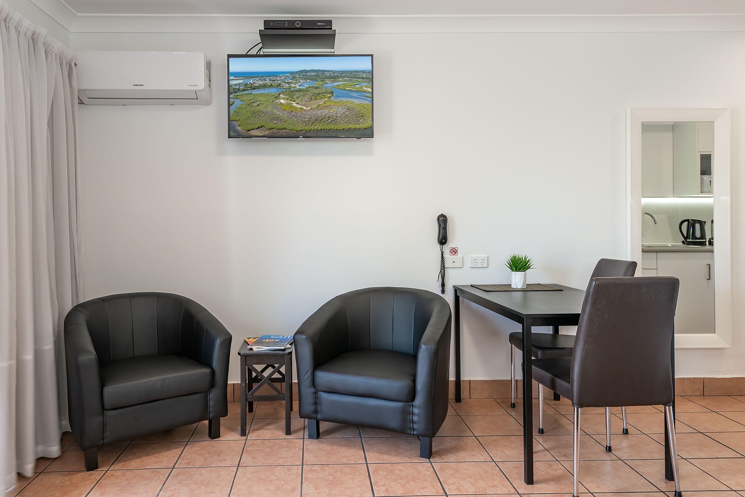 A modern room with two leather armchairs, a small table with a book, a flat-screen TV on the wall, and a kitchen area visible through an open door.