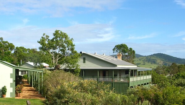 A green house with a porch and a pathway leading to it, surrounded by greenery and hills in the background. - Amamoor Lodge (Amamoor)
