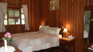 A wooden-paneled room with a bed, a nightstand, a bedside table, a vase with flowers, and a painting on the wall. - Amamoor Lodge (Amamoor)