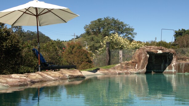 A clear swimming pool with a rock formation and a bench under a white umbrella. - Amamoor Lodge (Amamoor)