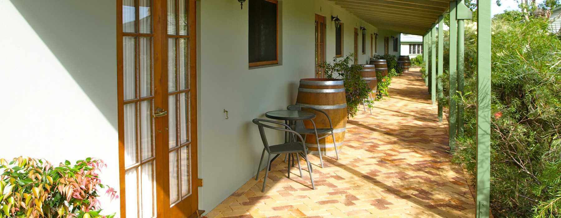 A covered outdoor area with a brick floor, wooden barrels, metal chairs, and a row of windows.