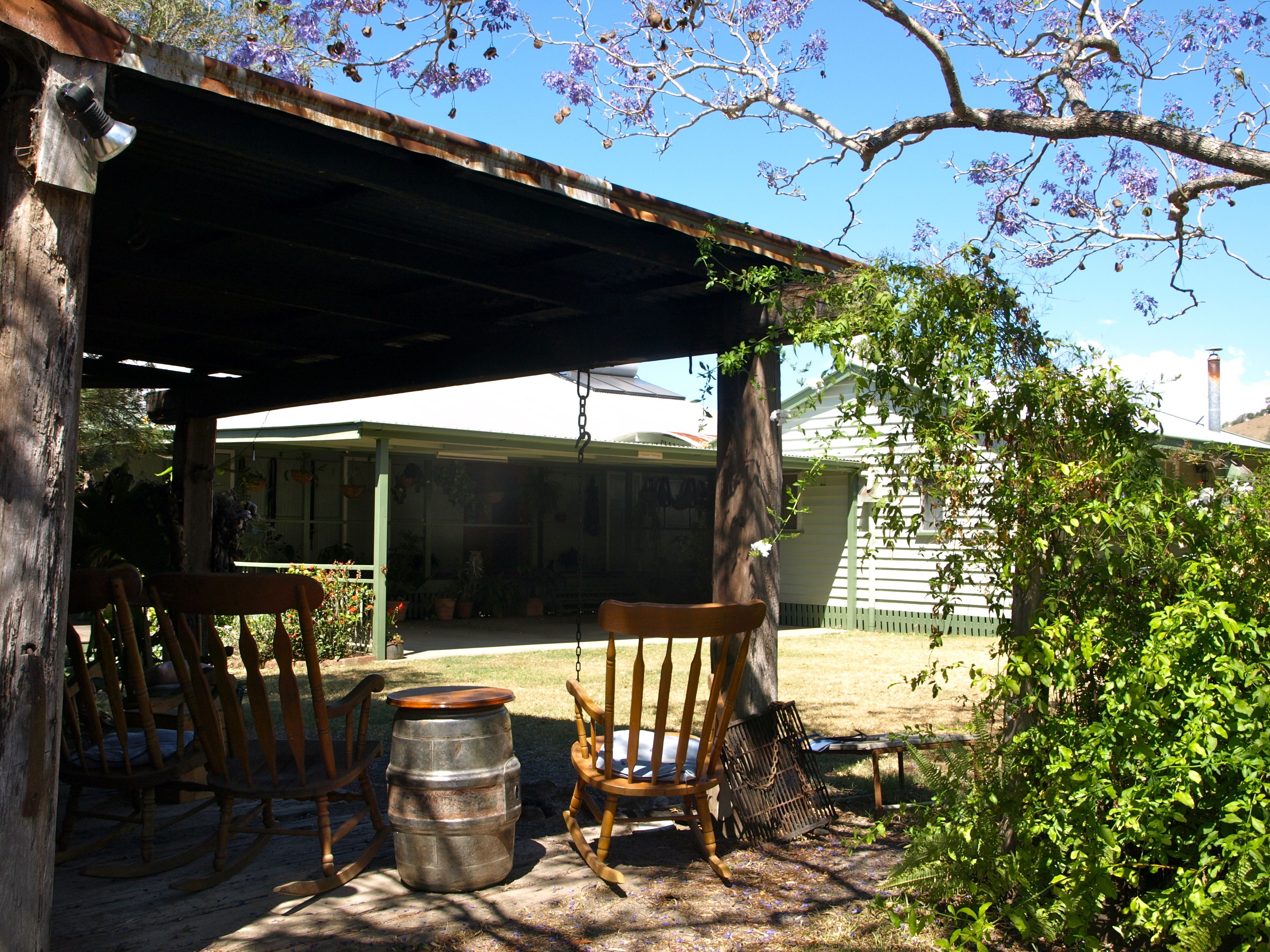 Un espace extérieur rustique aménagé avec des chaises en bois et un tonneau en métal, sous un toit métallique, entouré de verdure et avec une maison en arrière-plan.