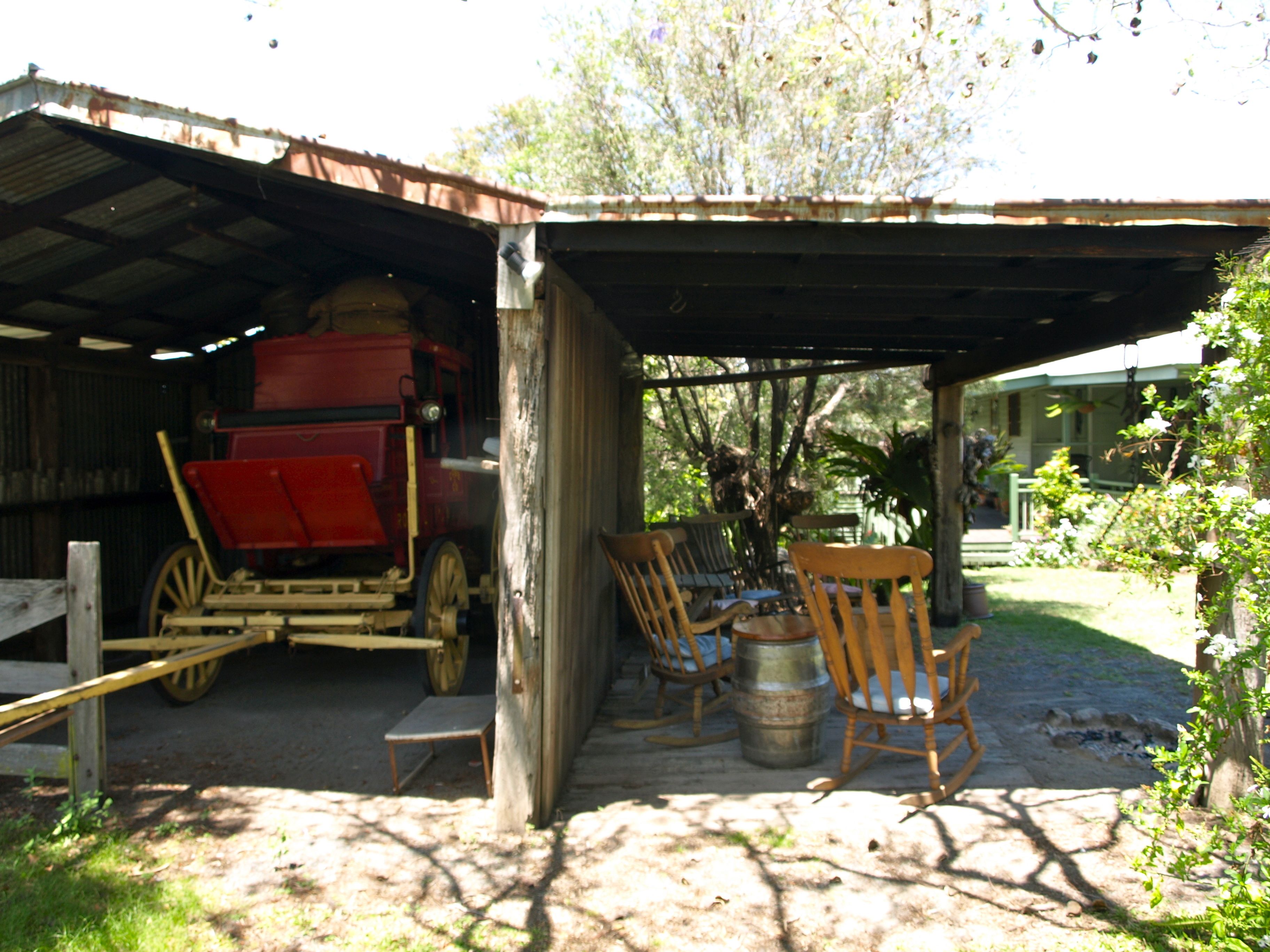 A rustic outdoor seating area with wooden chairs and a table, a red cart, and a covered structure with a metal roof.