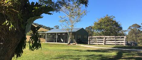 A green building with a covered outdoor area, surrounded by trees and a grassy field.