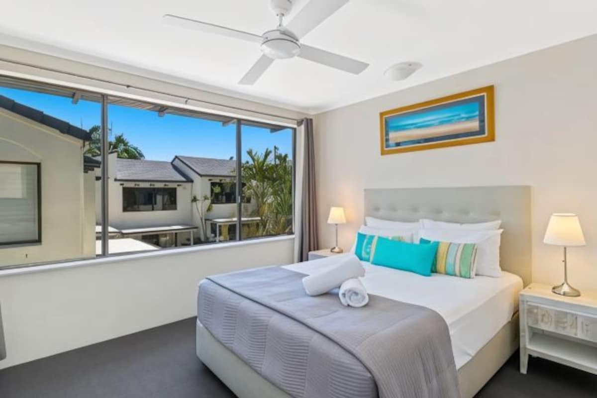 Habitación de hotel con cama, ventilador de techo, ventana con vistas y un cuadro en la pared.