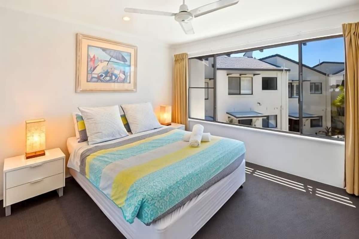 A hotel room with a bed, bedside lamps, a ceiling fan, a window with a view of neighboring houses, and a framed picture on the wall.