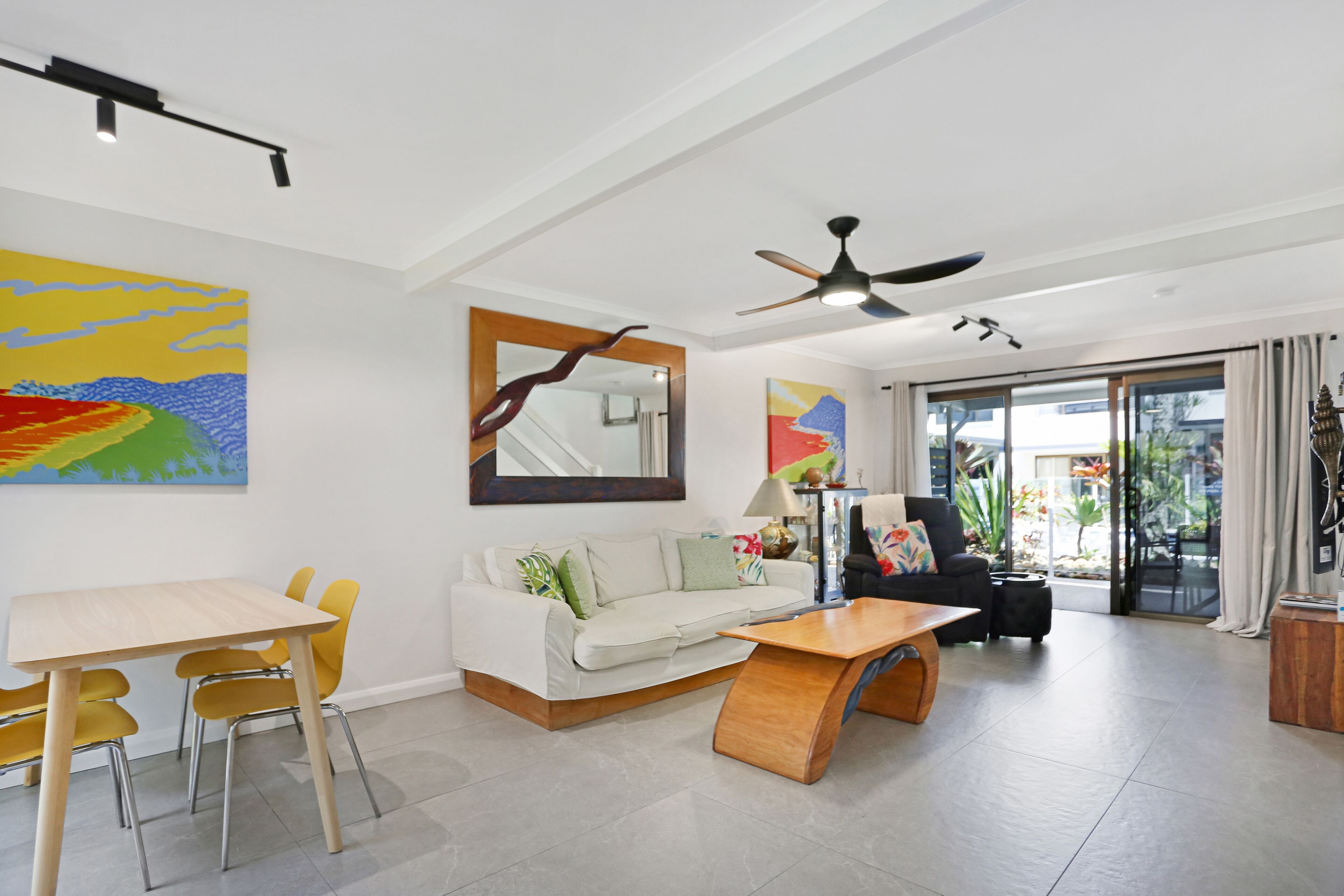 A modern living room with a sofa, dining table, and chairs, a ceiling fan, and a large window with a view of the outdoors.