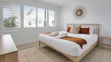 A bedroom with a bed, a bedside table, a nightstand, a window with blinds, and a wall-mounted mirror.
