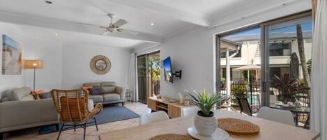 A modern living room with a sofa, a dining table with placemats, a ceiling fan, and a large window offering a view of the outdoors.