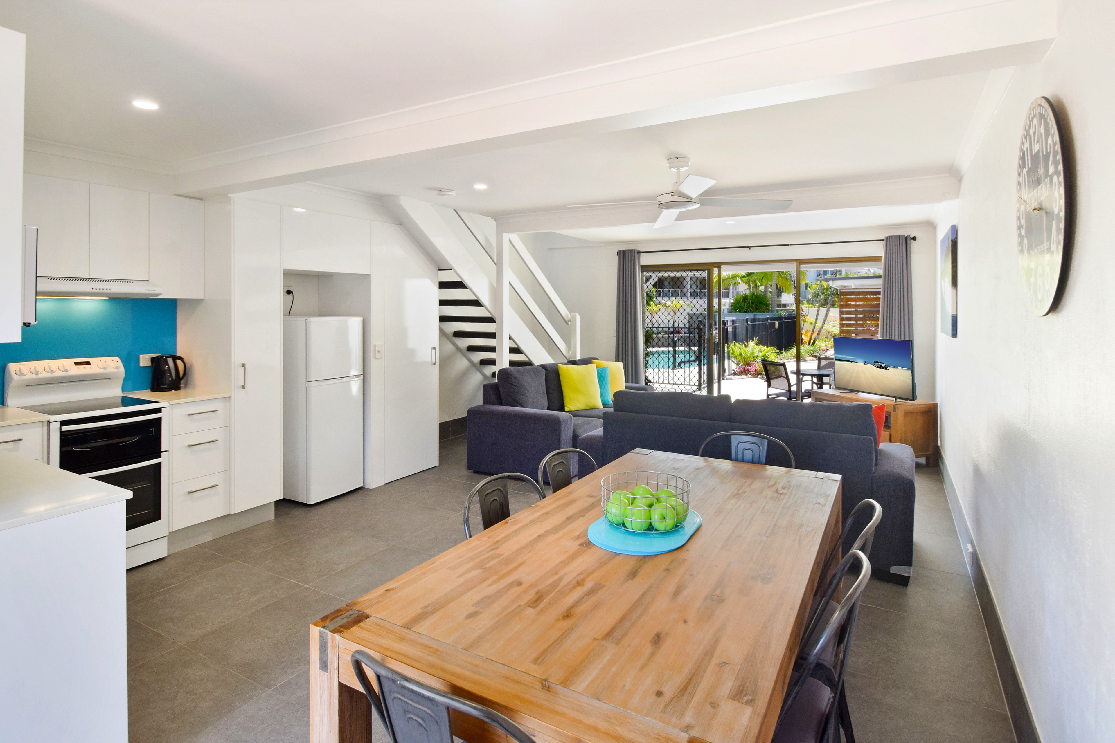 Une cuisine moderne avec des meubles blancs, une table à manger en bois sur laquelle est posé un panier de fruits, un escalier menant à un balcon et une vue sur une piscine.