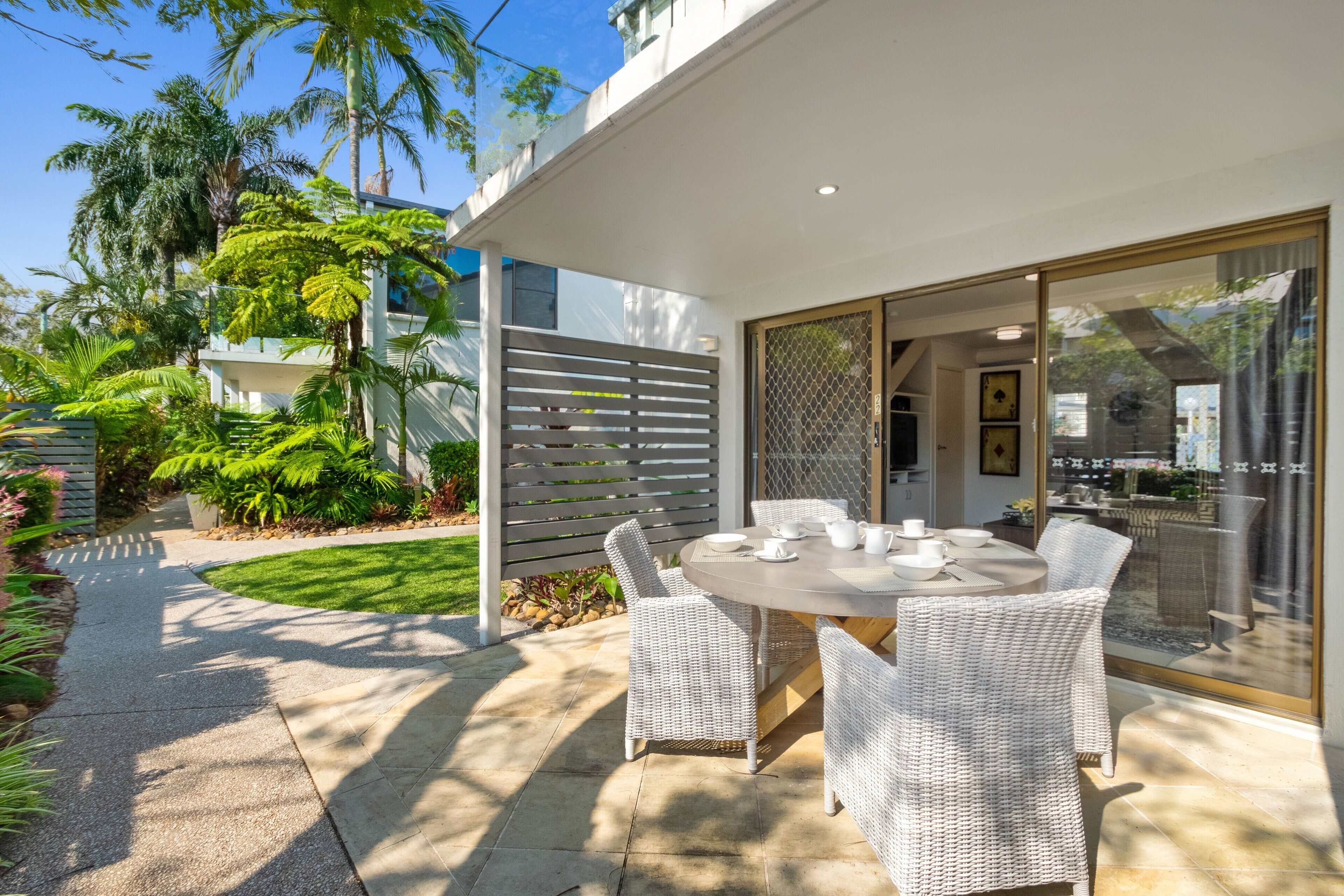 A patio with a round table and white wicker chairs, surrounded by lush greenery and a white fence.