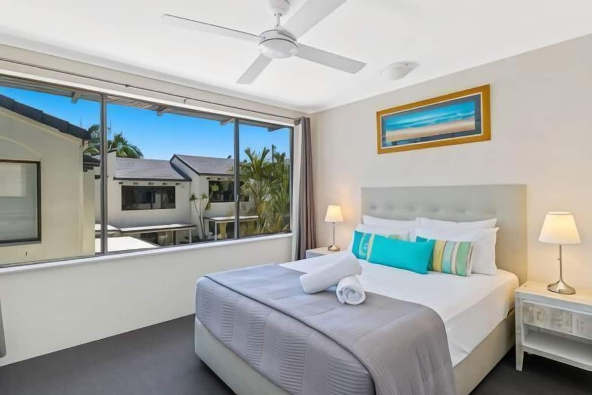 A hotel room with a bed, a ceiling fan, a window with a view, and a painting on the wall.