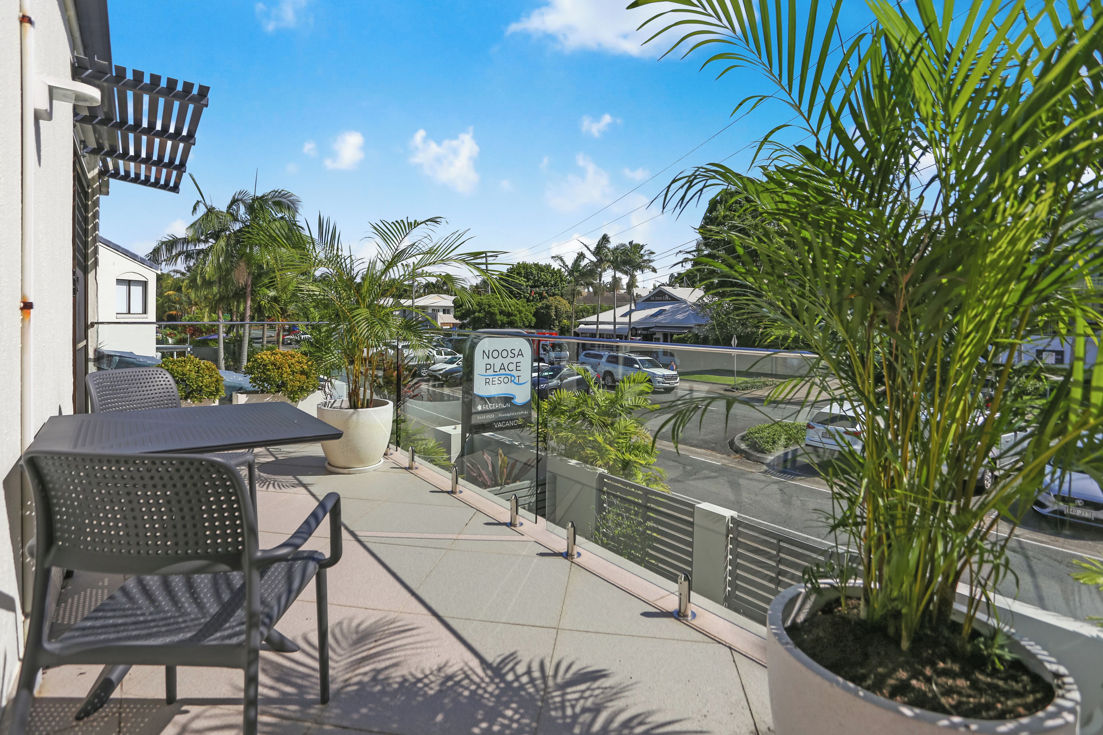 A balcony with outdoor furniture, a sign for Noosa Place Resort, and a view of the street with cars and palm trees.
