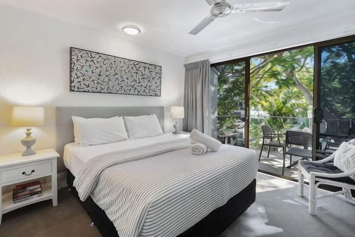 A bedroom with a bed, bedside tables, a ceiling fan, a large window, and a balcony with chairs.
