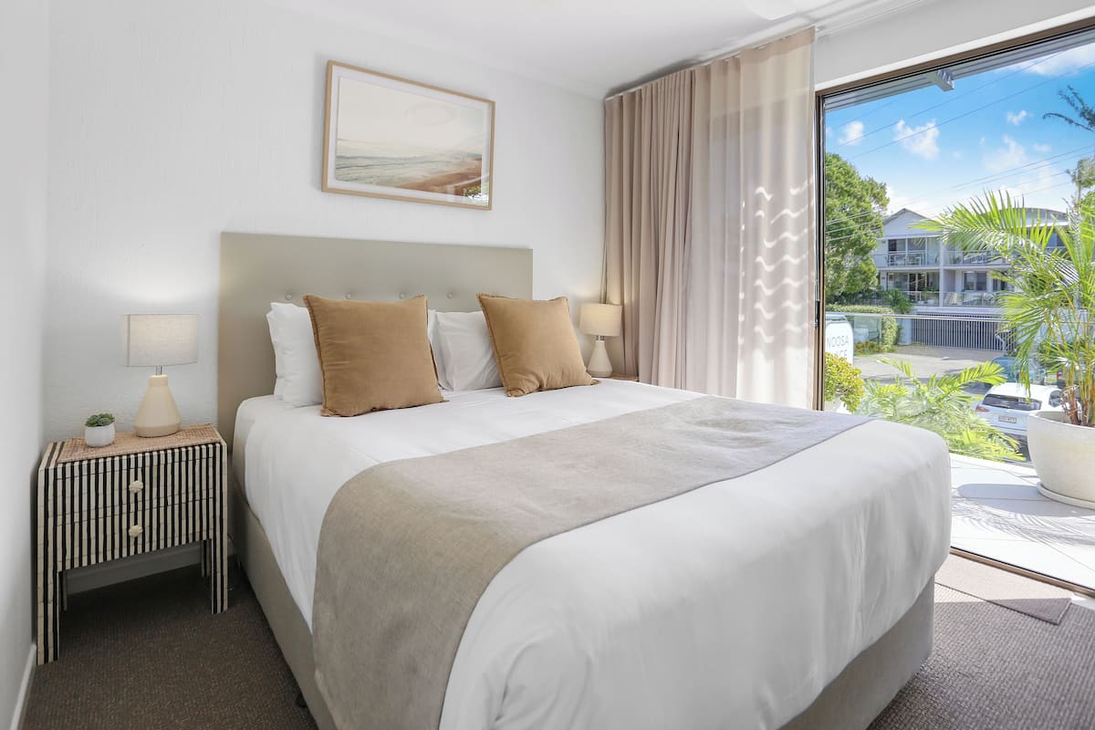 A hotel room with a large bed, a bedside table with a lamp, a painting on the wall, and a view of the outside.
