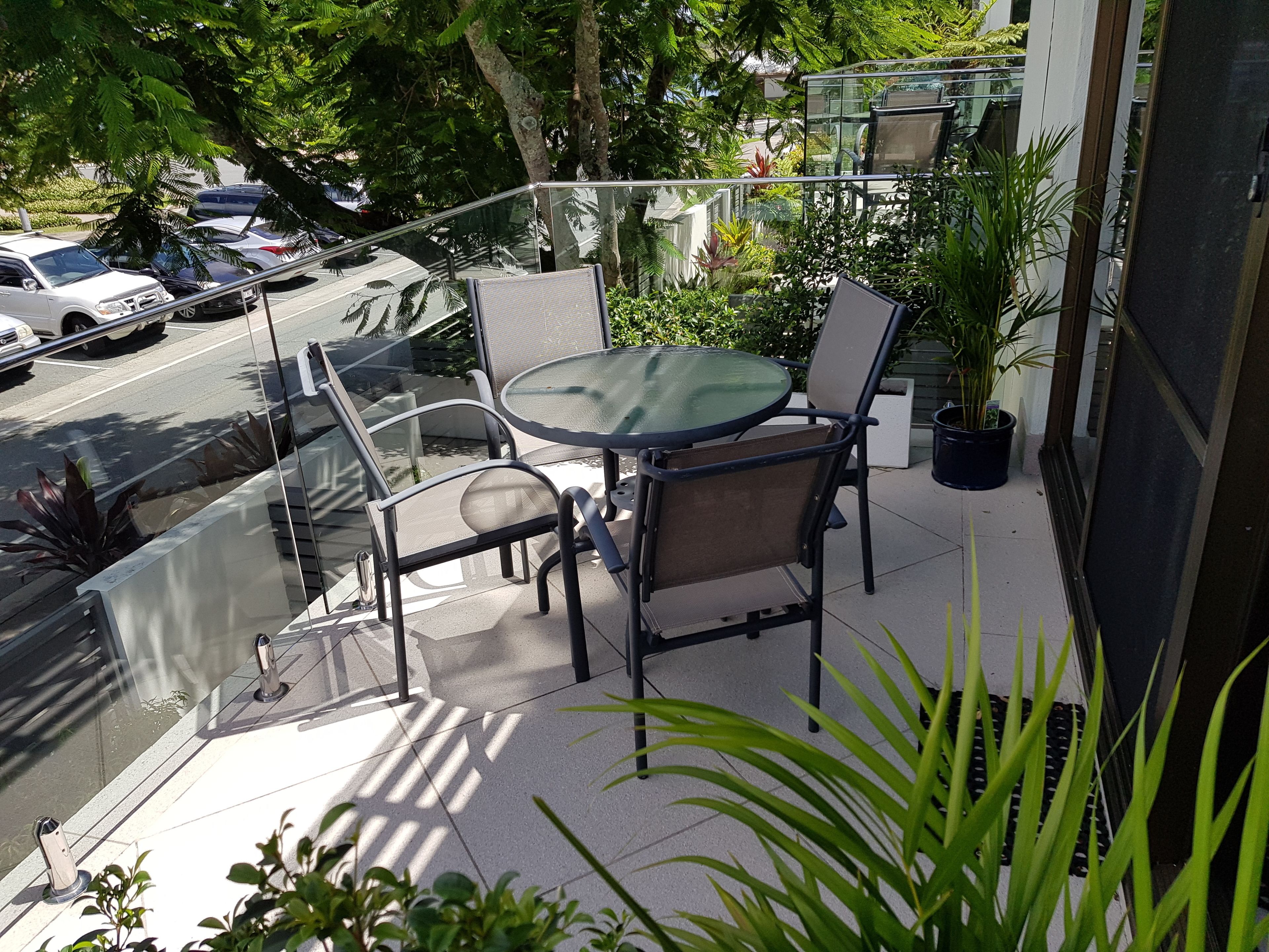 A balcony with a glass table and chairs, surrounded by potted plants and overlooking a parking area.