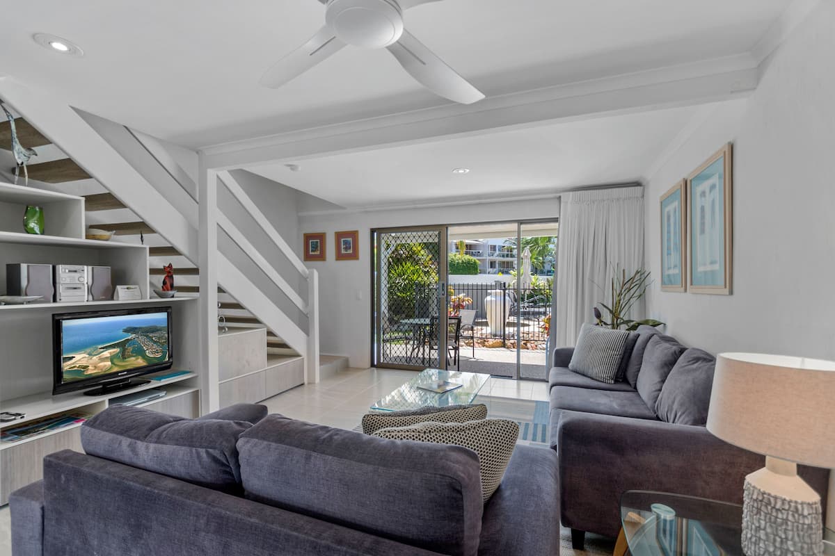 A modern living room with a flat-screen TV, grey sofas, a glass coffee table, and a ceiling fan.