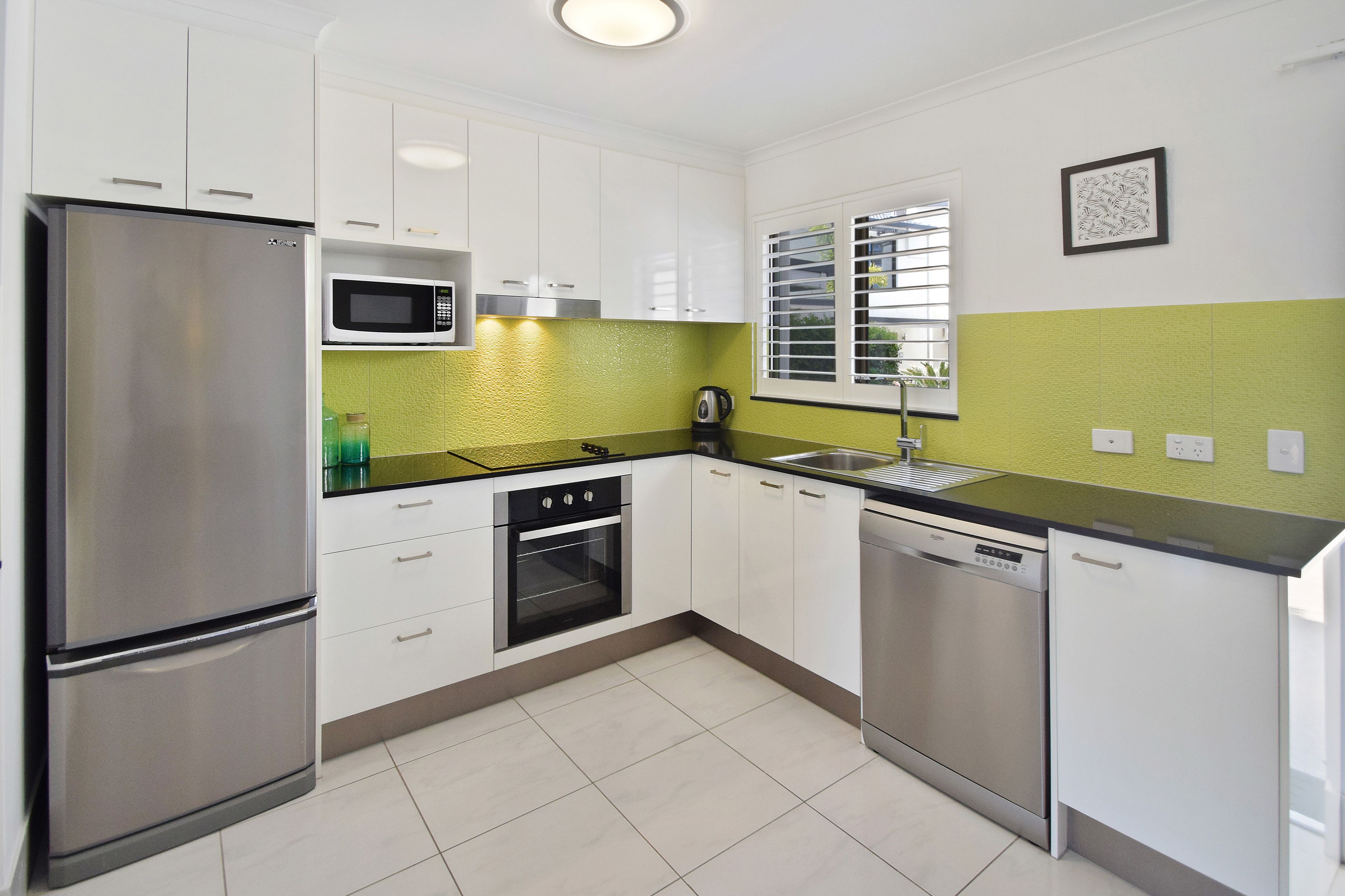 A modern kitchen with white cabinets, stainless steel appliances, a green backsplash, and a window with blinds.
