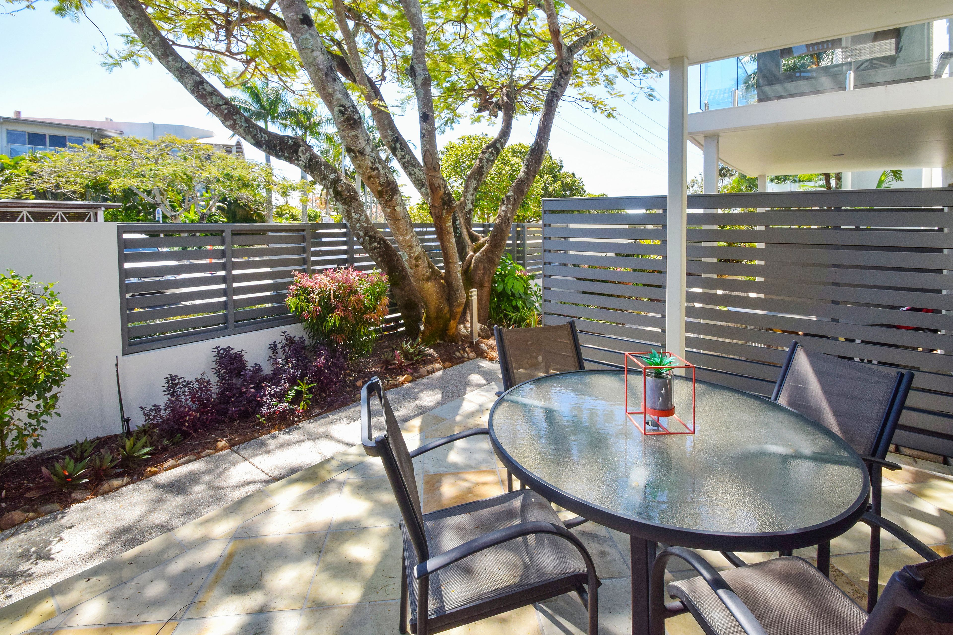 An outdoor patio with a round glass table and four chairs, surrounded by a white fence and various plants.